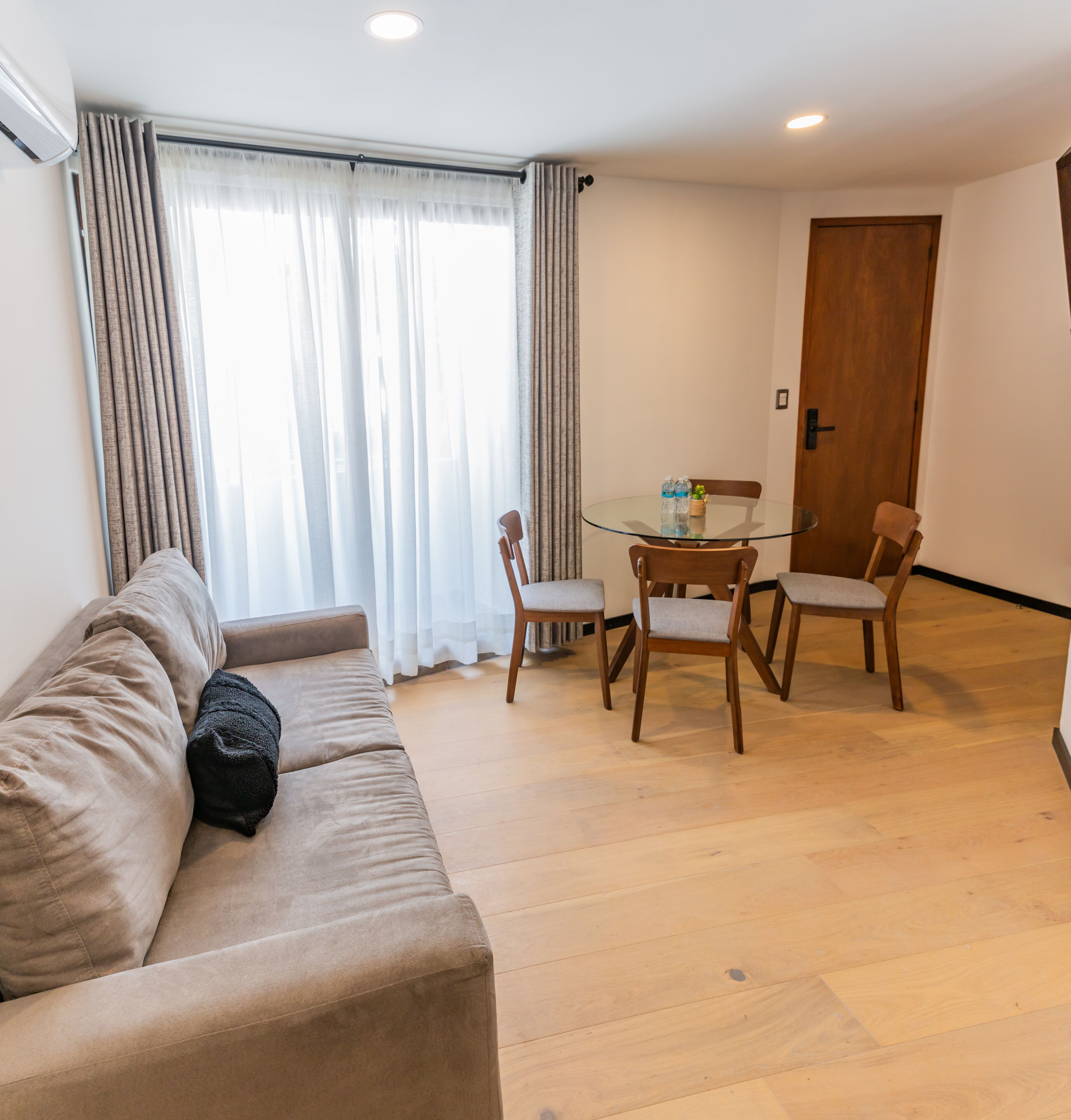 A living room with a beige sofa, a glass-top dining table with four wooden chairs, and a wooden door.