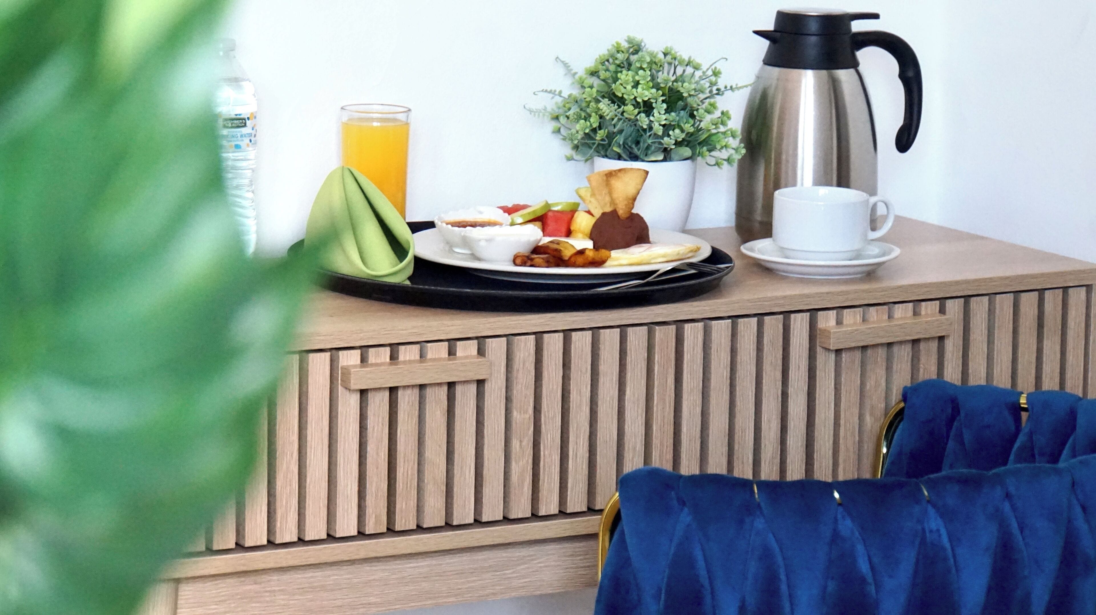 Una bandeja de desayuno con comida y bebidas sobre una encimera de madera, una cafetera, una taza blanca y una pequeña planta.