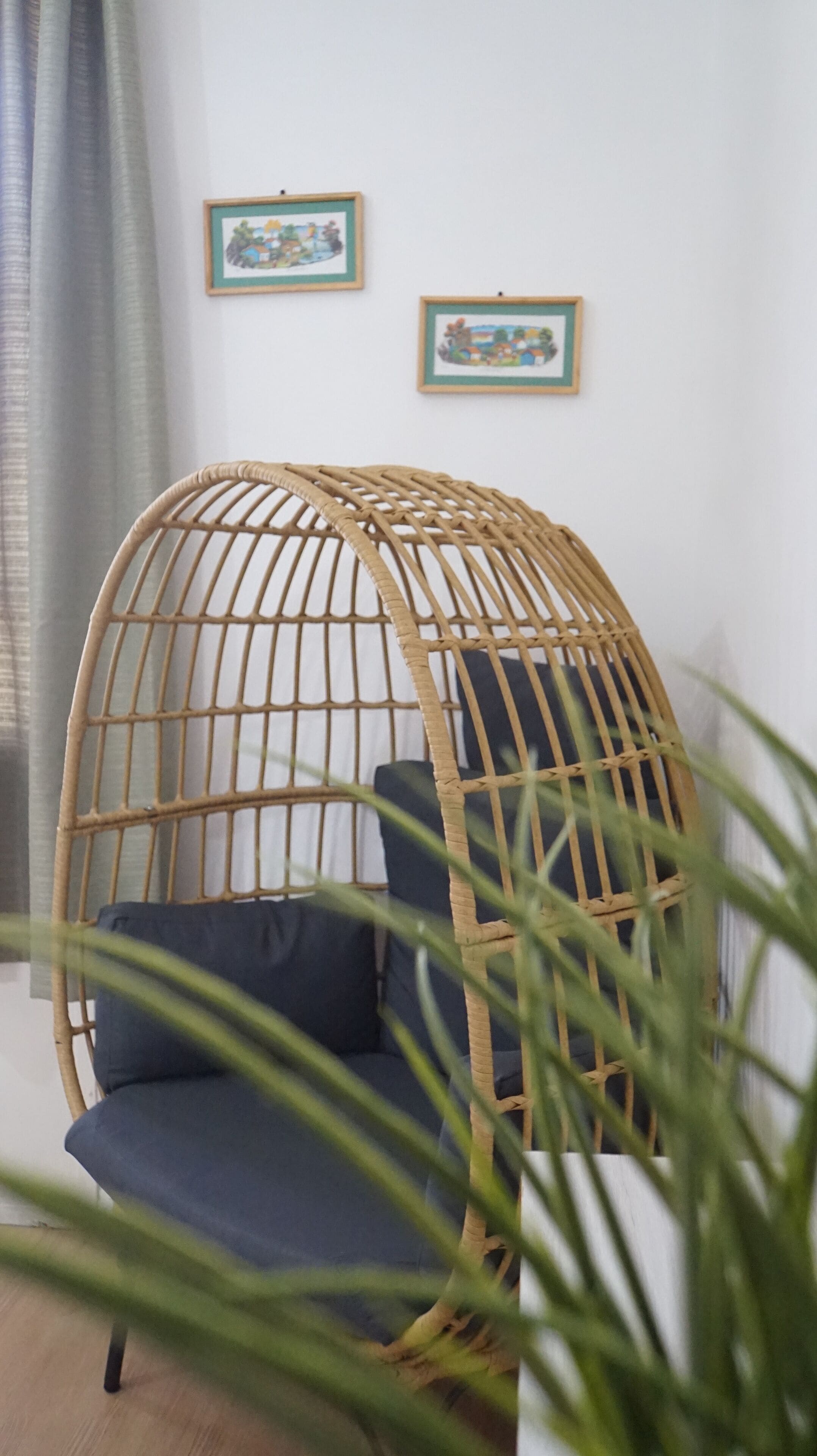 A wicker hanging chair with a cushion, a grey curtain, and two framed pictures on the wall.