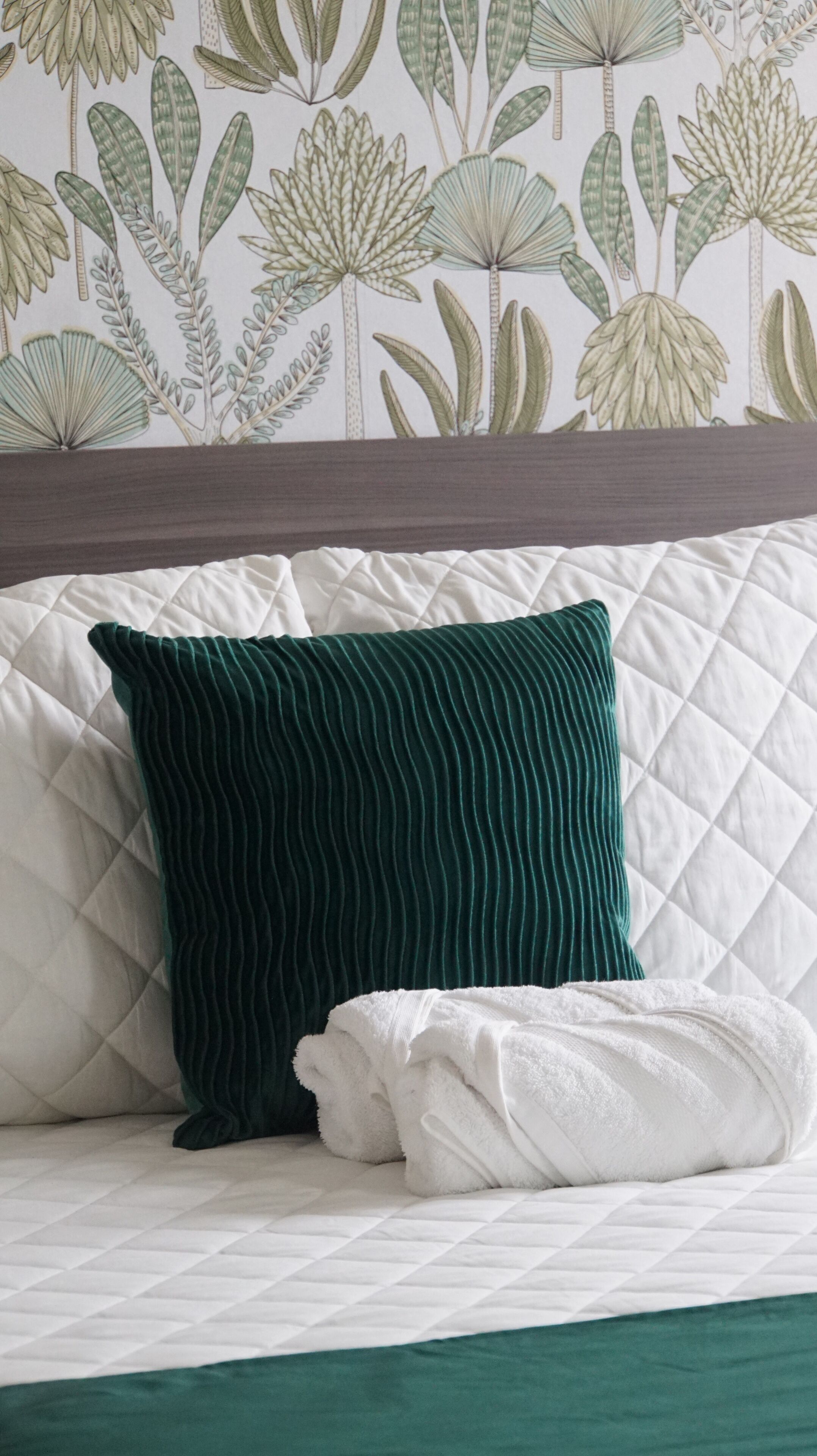 A bed with a quilted white cover, a green ribbed pillow, and a white towel.