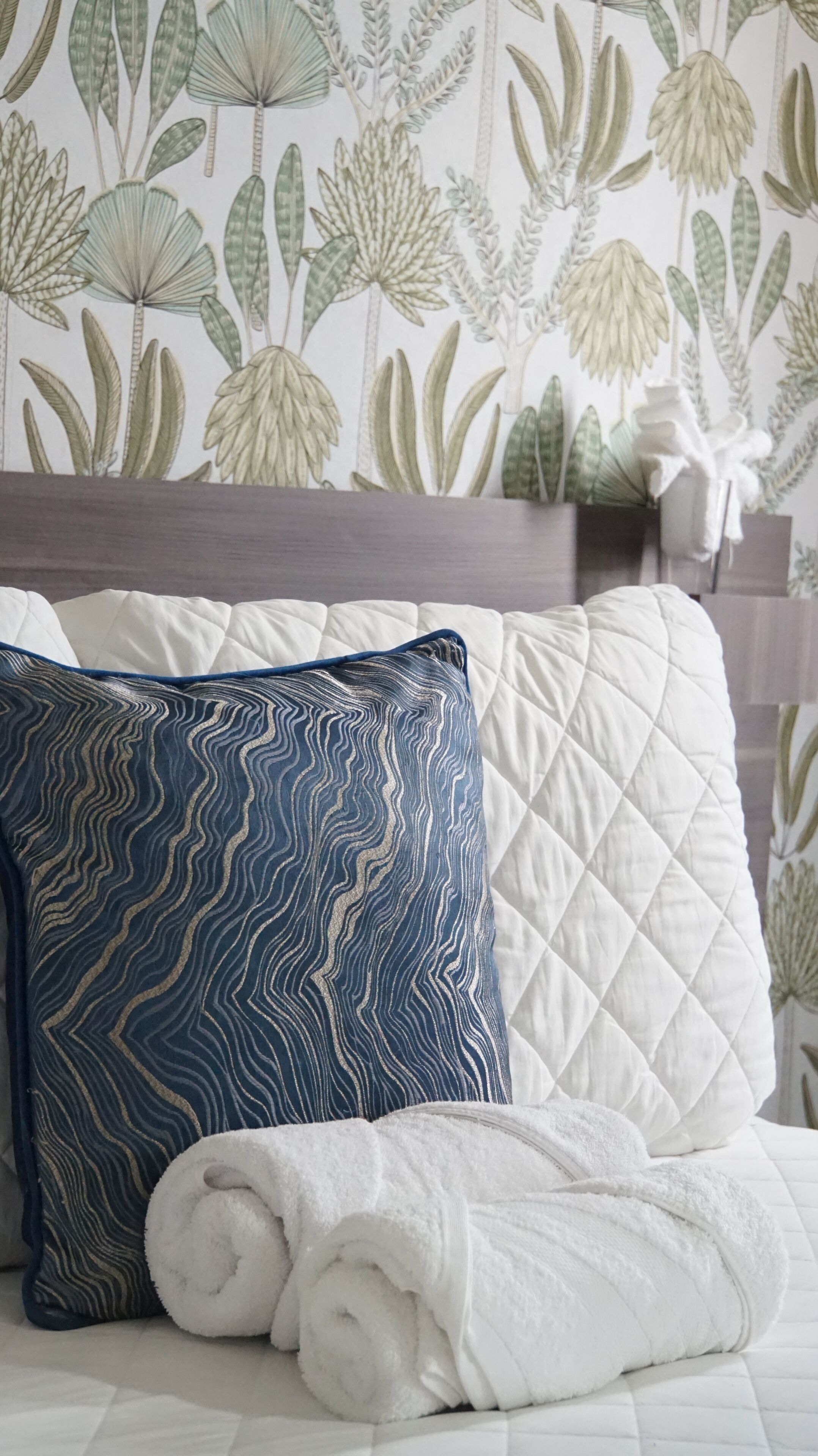 Cama bem arrumada com edredom branco acolchoado, toalha branca enrolada e travesseiro azul com padrão ondulado.