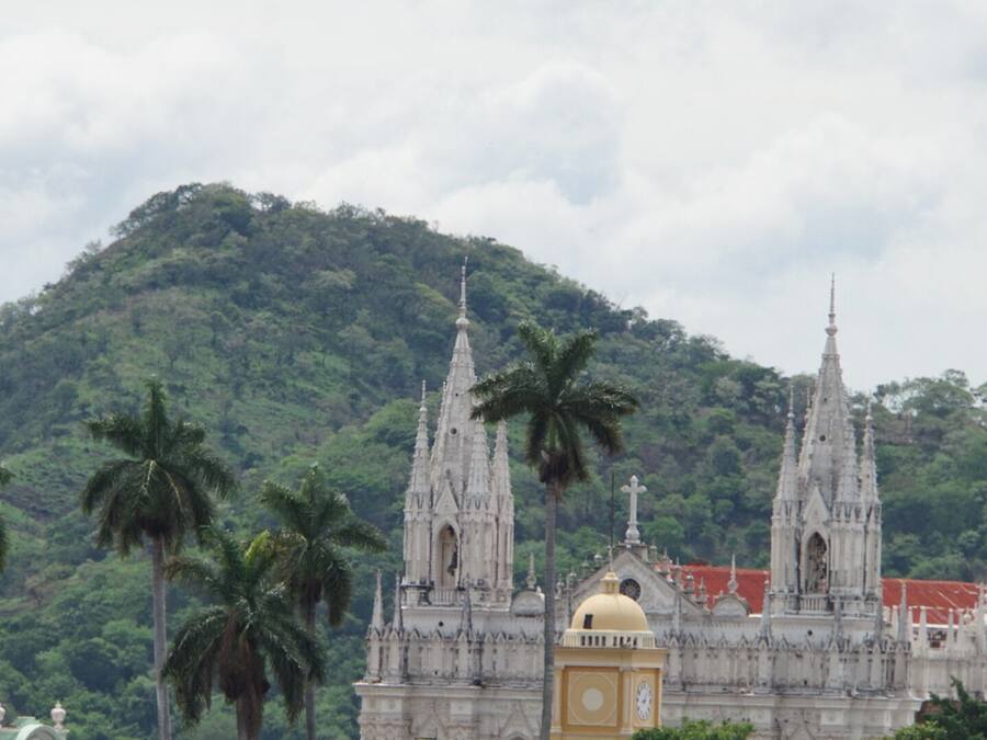 A historic building with two tall spires and a central dome, surrounded by lush greenery and palm trees.