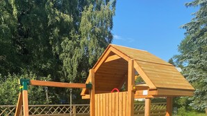 A wooden play structure with a slide and climbing wall, surrounded by a fence and grass. - Cozy holiday home in the heart of the Ore Mountains. (Sehmatal)