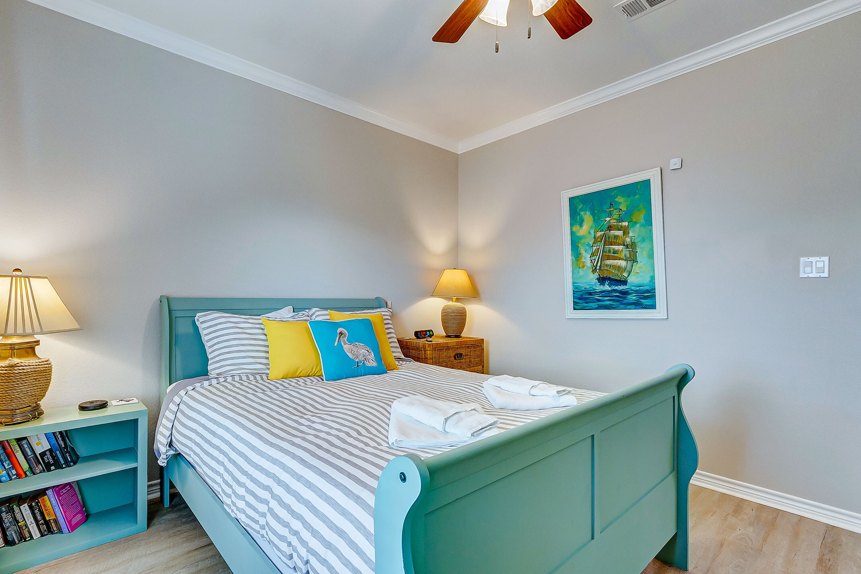 A bedroom with a teal bed, striped bedding, a nightstand with a lamp, a bookshelf, and a painting of a ship on the wall.