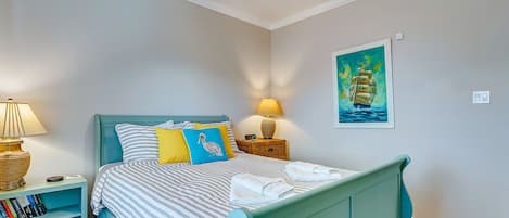 A bedroom with a teal bed, striped bedding, a nightstand with a lamp, a bookshelf, and a painting of a ship on the wall.