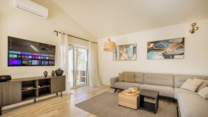 Ein modernes Wohnzimmer mit grauem Sofa, Couchtisch und flachbildfernseher an der Wand. Darunter befindet sich eine Holzkommode und ein großes Fenster mit Vorhängen.