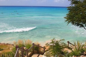 A beach with clear turquoise water, a wooden staircase leading down to the shore, and some greenery in the foreground.