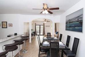 Un espace repas avec une longue table, des chaises noires et un ventilateur de plafond. Un comptoir de bar avec des tabourets se trouve à gauche.