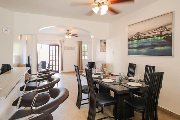 A dining area with a long table and chairs, a ceiling fan, and a large painting of a bridge.