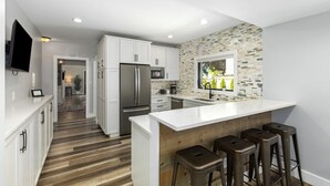 Una cocina moderna con armarios blancos, una isla grande con taburetes, un salpicadero de piedra e iluminación empotrada.