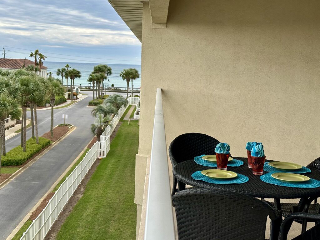 A balcony with two wicker chairs, a table set with plates and cups, overlooking a street lined with palm trees and a view of the ocean.