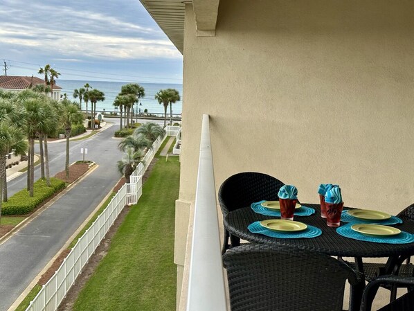 A balcony with two wicker chairs, a table set with plates and cups, overlooking a street lined with palm trees and a view of the ocean.