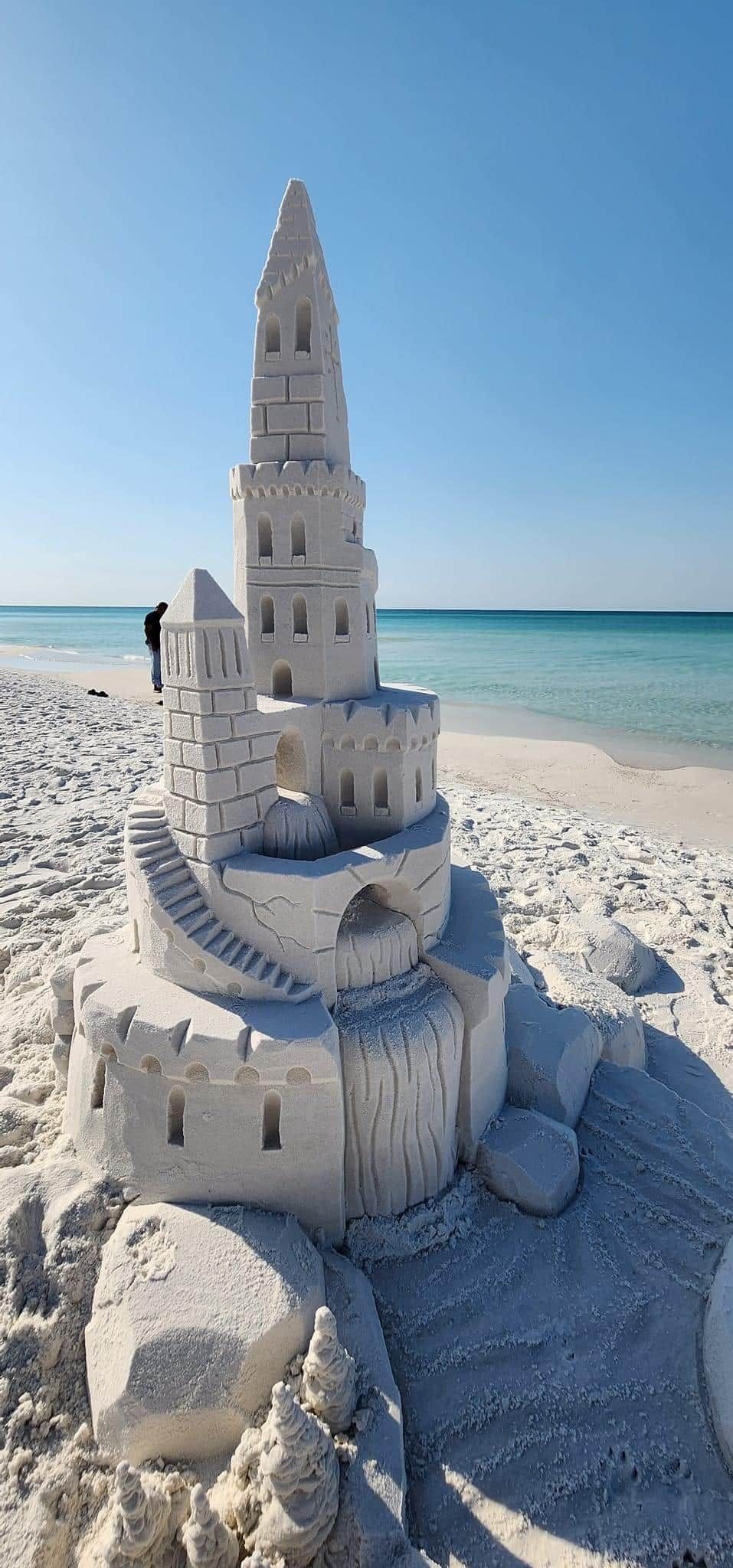 A sandcastle with multiple levels and a tower, situated on a sandy beach with a clear sky.