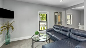 A modern living room with a leather sofa, a glass coffee table, a flat-screen TV mounted on the wall, and a view of the outside through a window. - Perfect 2-Bedroom Suite in Downtown Saugatuck! (Upper Level Unit) (Saugatuck)