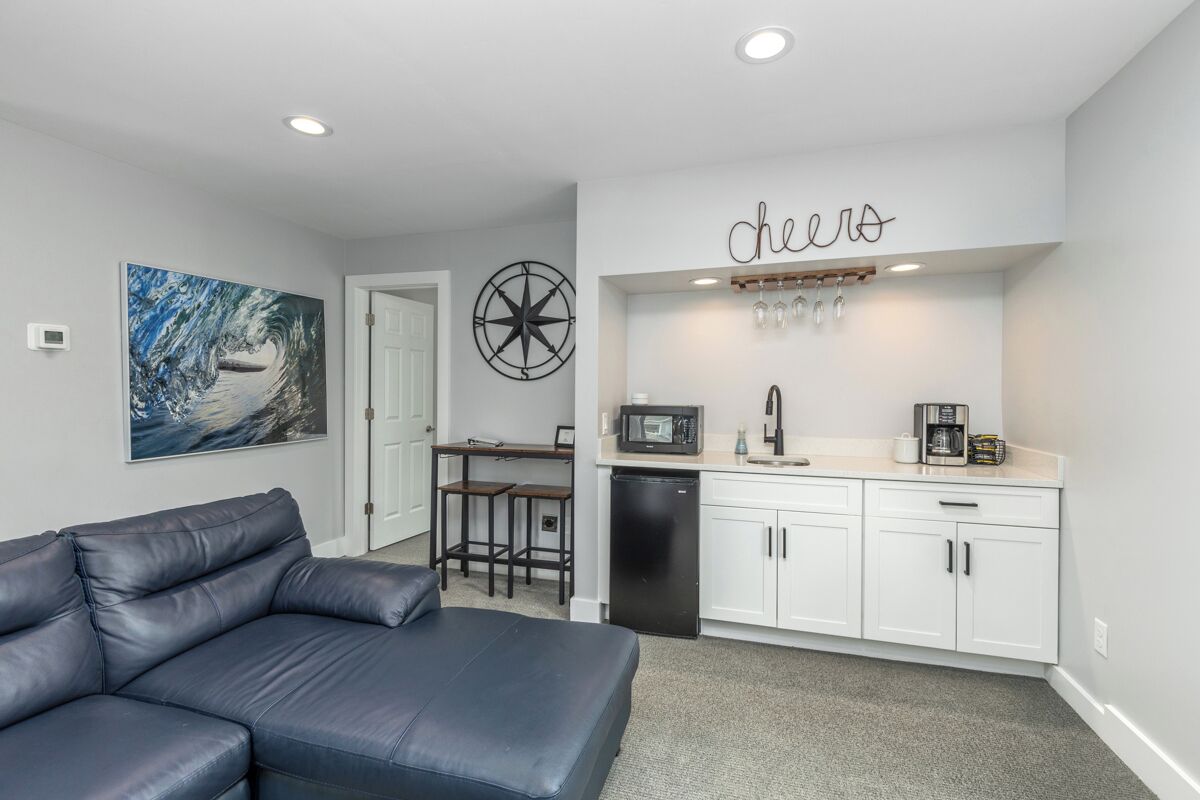 A modern kitchen with white cabinets, a black refrigerator, and a microwave. There is a seating area with a black leather couch and a small table. A large painting of a wave is on the wall, and a sign that says 'cheers' is above the kitchen counter.