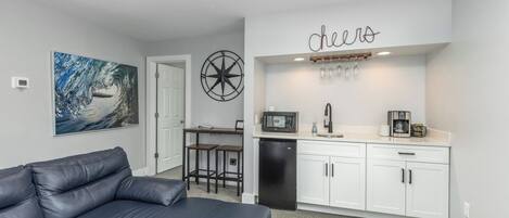 A modern kitchen with white cabinets, a black refrigerator, and a microwave. There is a seating area with a black leather couch and a small table. A large painting of a wave is on the wall, and a sign that says 'cheers' is above the kitchen counter.