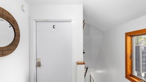 Une porte blanche avec la lettre « C » au-dessus, une fenêtre à cadre en bois et un miroir rond accroché au mur.