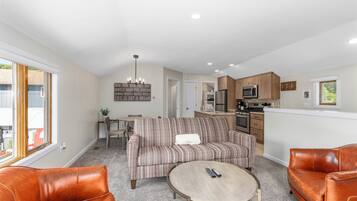 Een moderne woonkamer met een bank, salontafel en lederen fauteuils. In de achtergrond is een keuken met houten kastjes en apparatuur zichtbaar.
