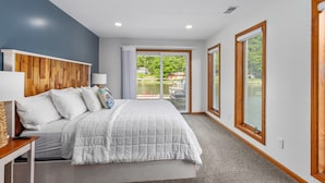 A bedroom with a bed, a nightstand, a lamp, a wooden headboard, a view of the outdoors, and a sliding glass door.