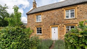 Une maison en pierre avec une porte blanche et deux fenêtres, entourée d’une végétation luxuriante.