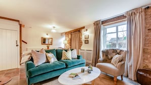 Un salon chaleureux avec un canapé de couleur turquoise, une table basse ronde et un fauteuil beige. On y trouve une grande fenêtre avec des rideaux, un mur en pierre et une porte dotée d’une poignée en bois.