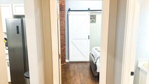 Un couloir doté d’un plancher en bois, une porte blanche donnant sur une pièce où se trouve un lit, et un réfrigérateur dans la pièce adjacente.