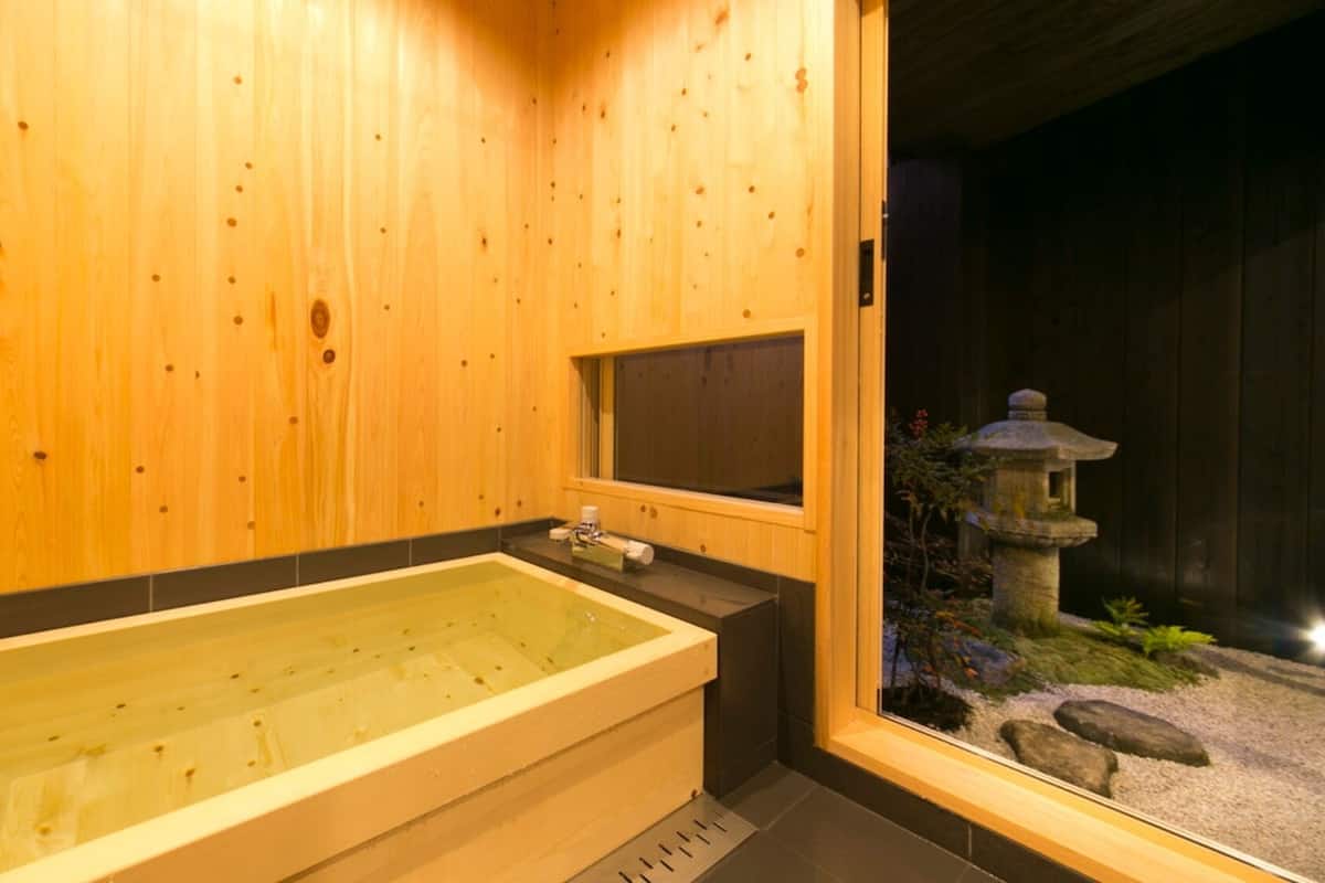 A wooden spa room with a large bathtub, a small window, and a stone lantern outside.