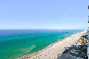 En strand med en mole, der strækker sig ud i havet, klare himmel og en sandstrand med parasoller.