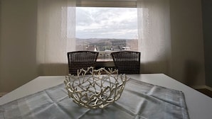 A dining area with a table set for two, a decorative metal bowl centerpiece, and a view of a landscape through a window with sheer curtains.