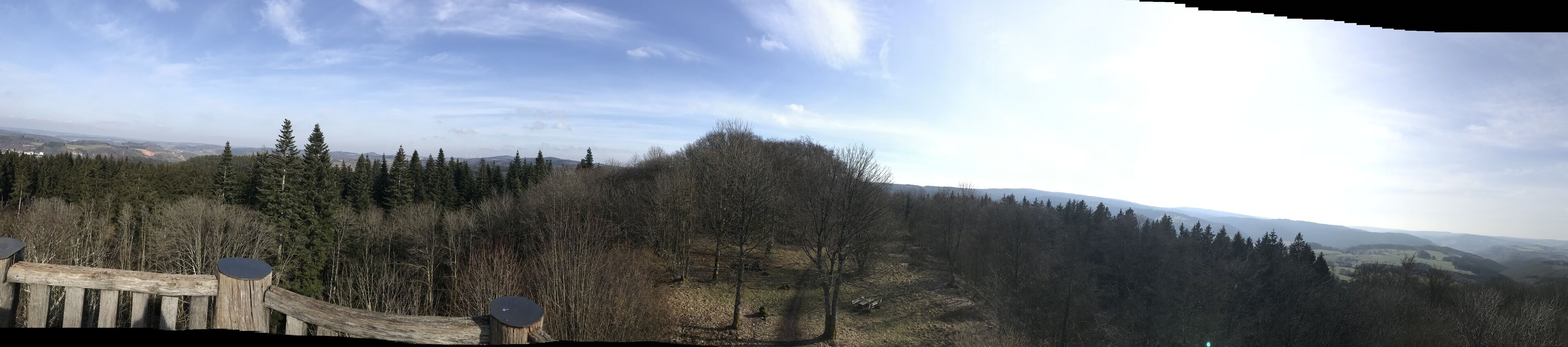 A panoramic view of a forested landscape with a wooden fence and a circular wooden post.
