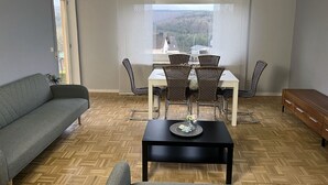 A modern living room with a sofa, a dining table with chairs, a coffee table, and a view of a hilly landscape through a window.