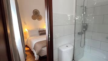 A bathroom with a glass-enclosed shower, a white toilet, and a bed in the adjacent room.