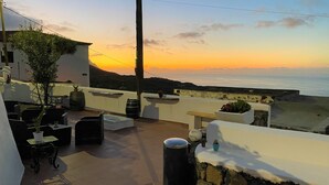 An outdoor terrace with seating, a view of the sea, and a sunset.
