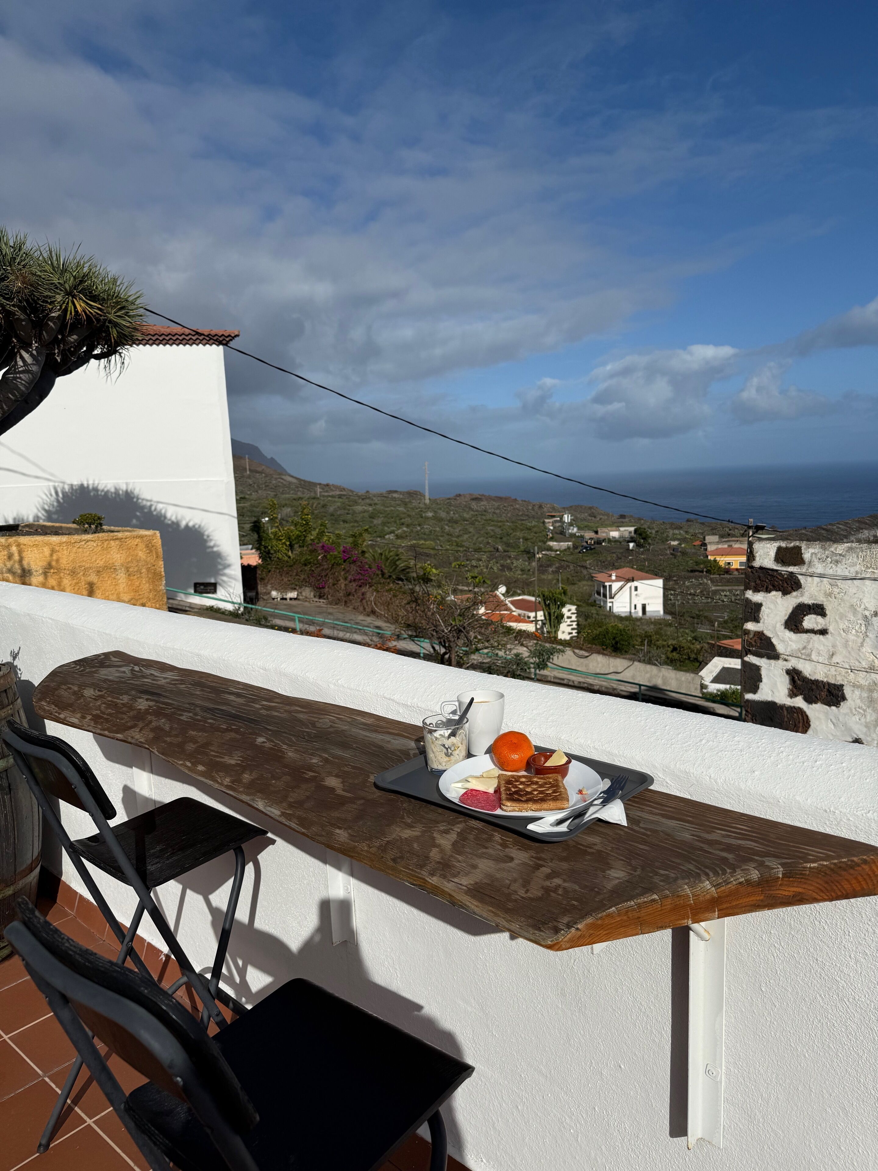 Un balcón con una mesa dispuesta para el desayuno, con vistas a un paisaje costero con casas y el mar.