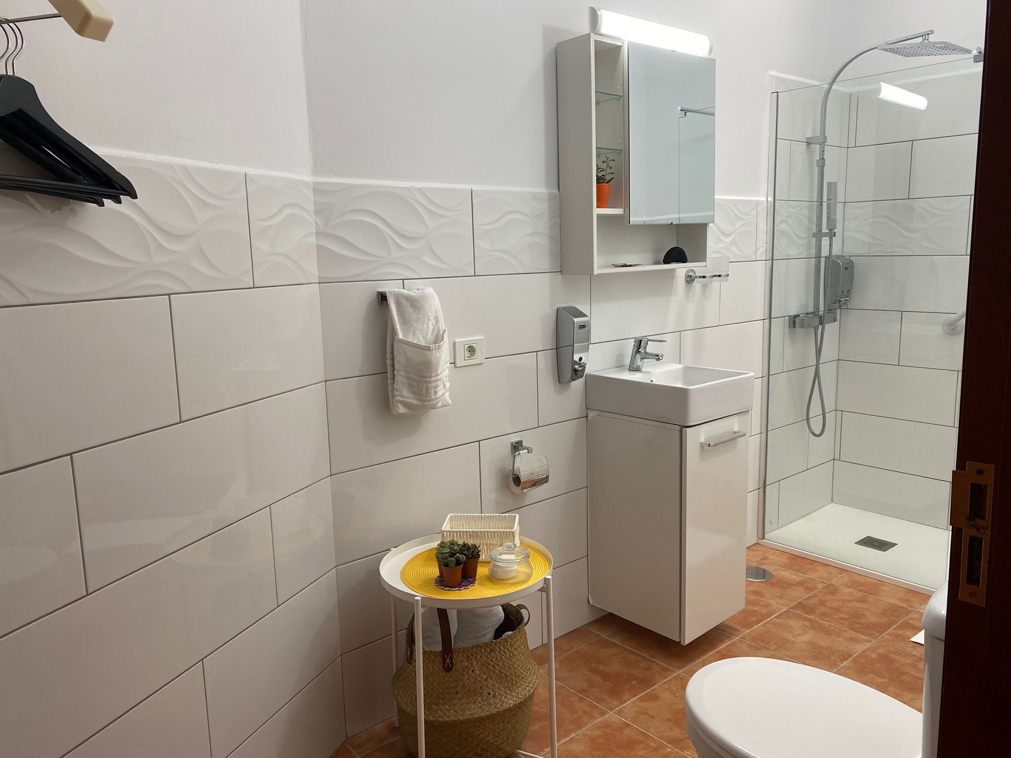 A bathroom with white tiles, a sink, a mirror, a towel rack, a small table with a plant, and a shower area.