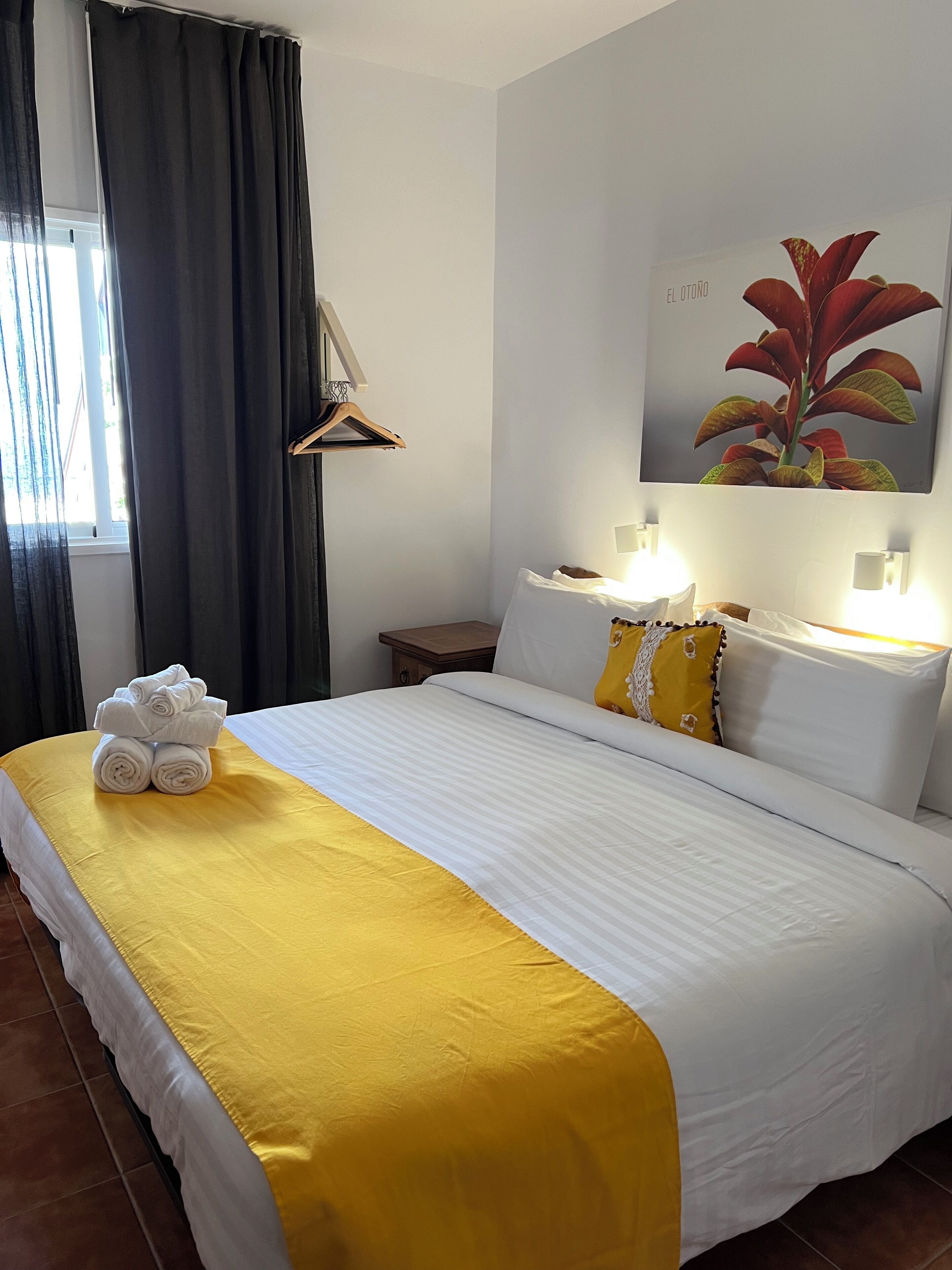 Cama bem arrumada com colcha amarela e branca, uma pilha de toalhas brancas e um quadro com folhas vermelhas na parede.