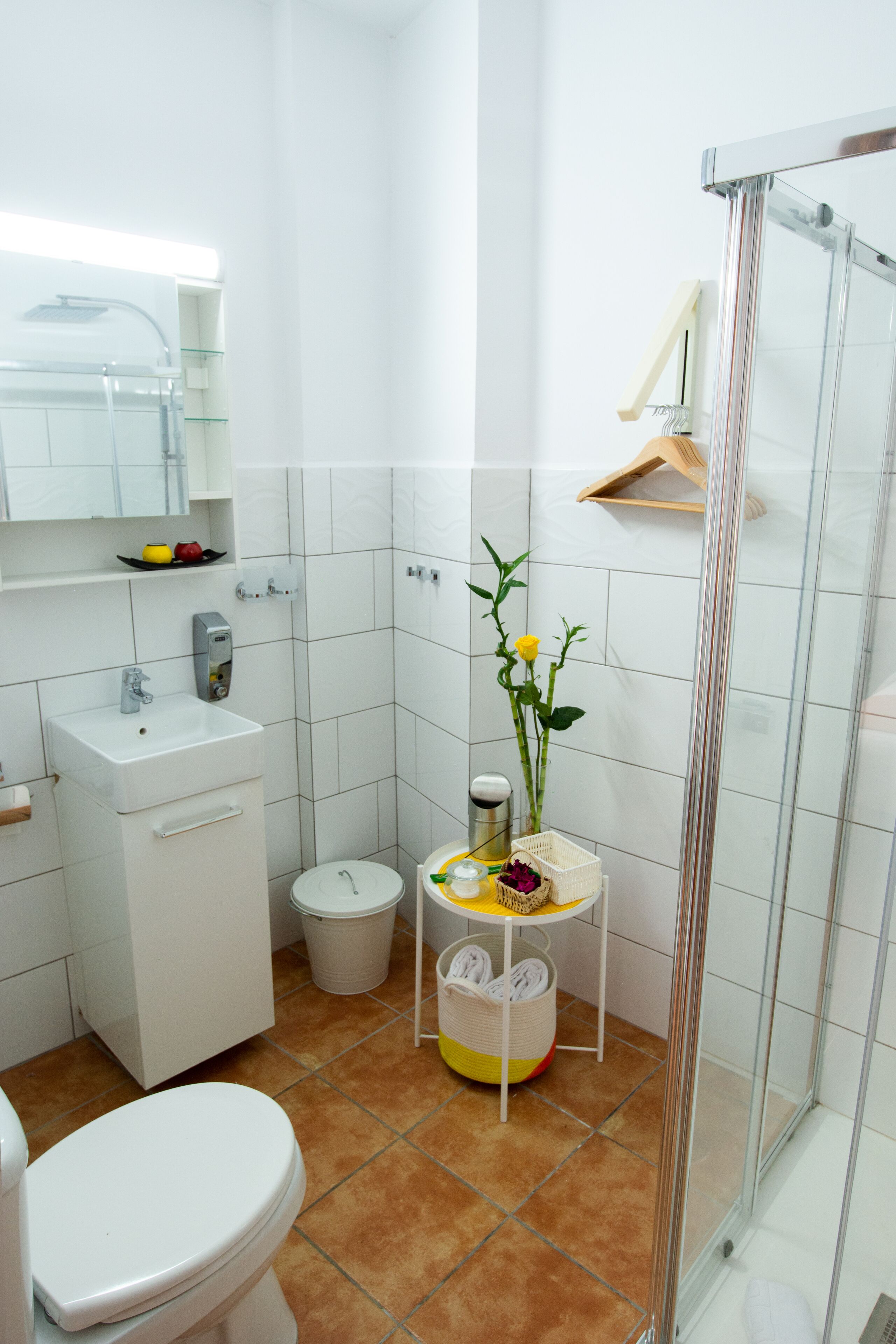 A bathroom with a white sink, toilet, and shower. There is a small plant on a shelf and a basket with towels.