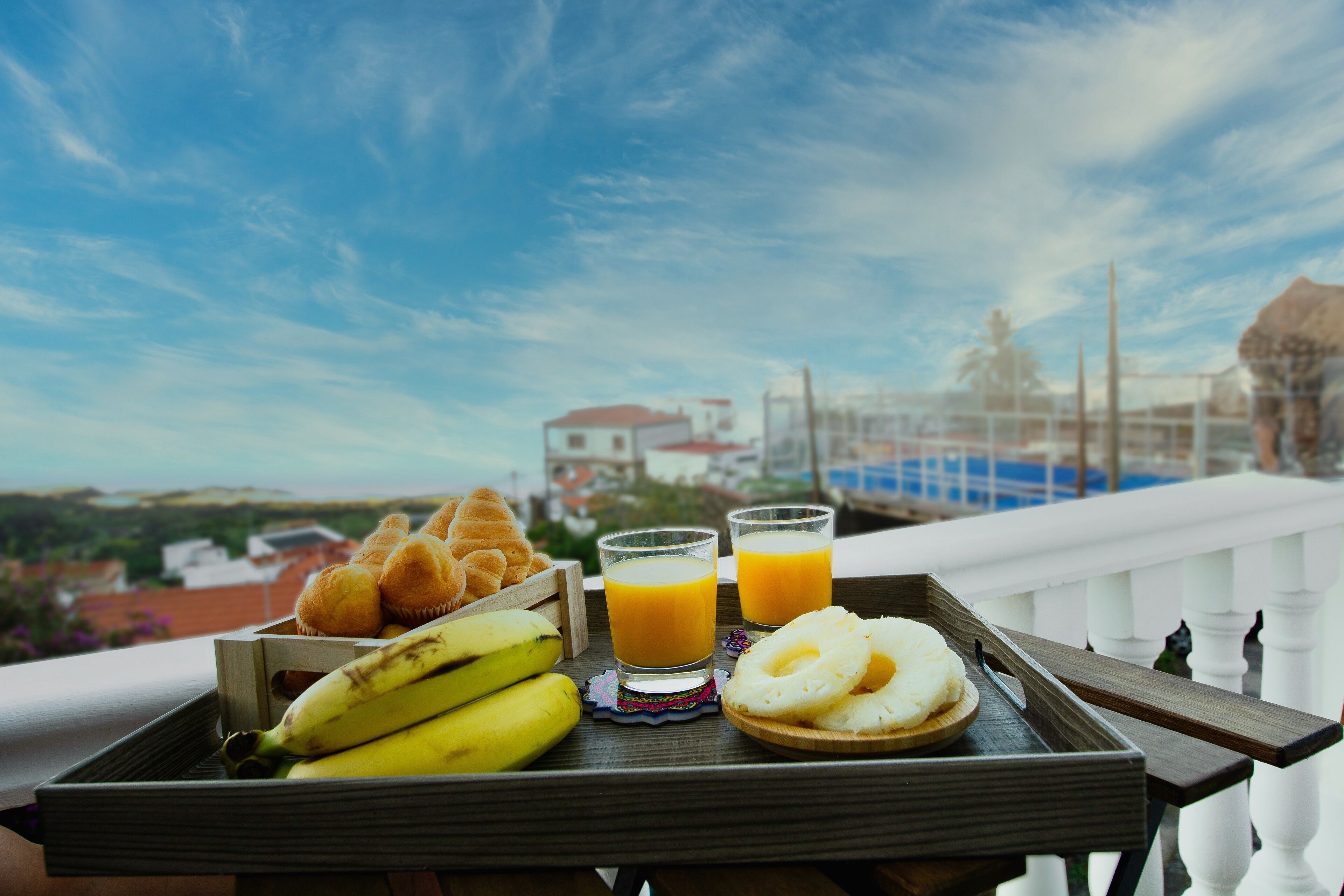A tray with bananas, muffins, and glasses of orange juice on a balcony with a view of buildings and a clear sky.
