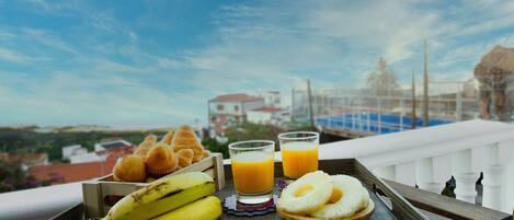 A tray with bananas, muffins, and glasses of orange juice on a balcony with a view of buildings and a clear sky.