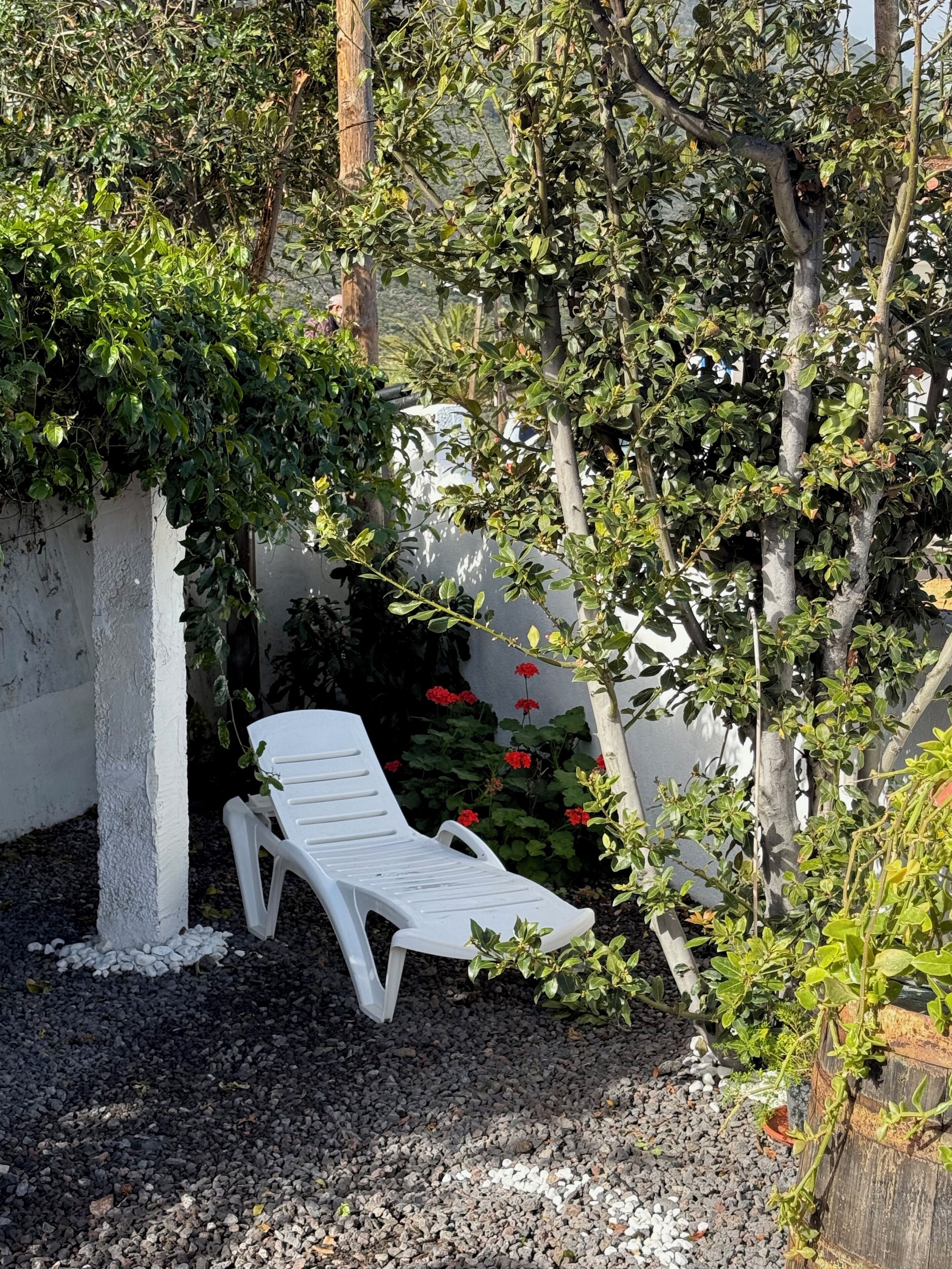 A white plastic chair in a garden area with gravel ground, surrounded by greenery and a white wall.