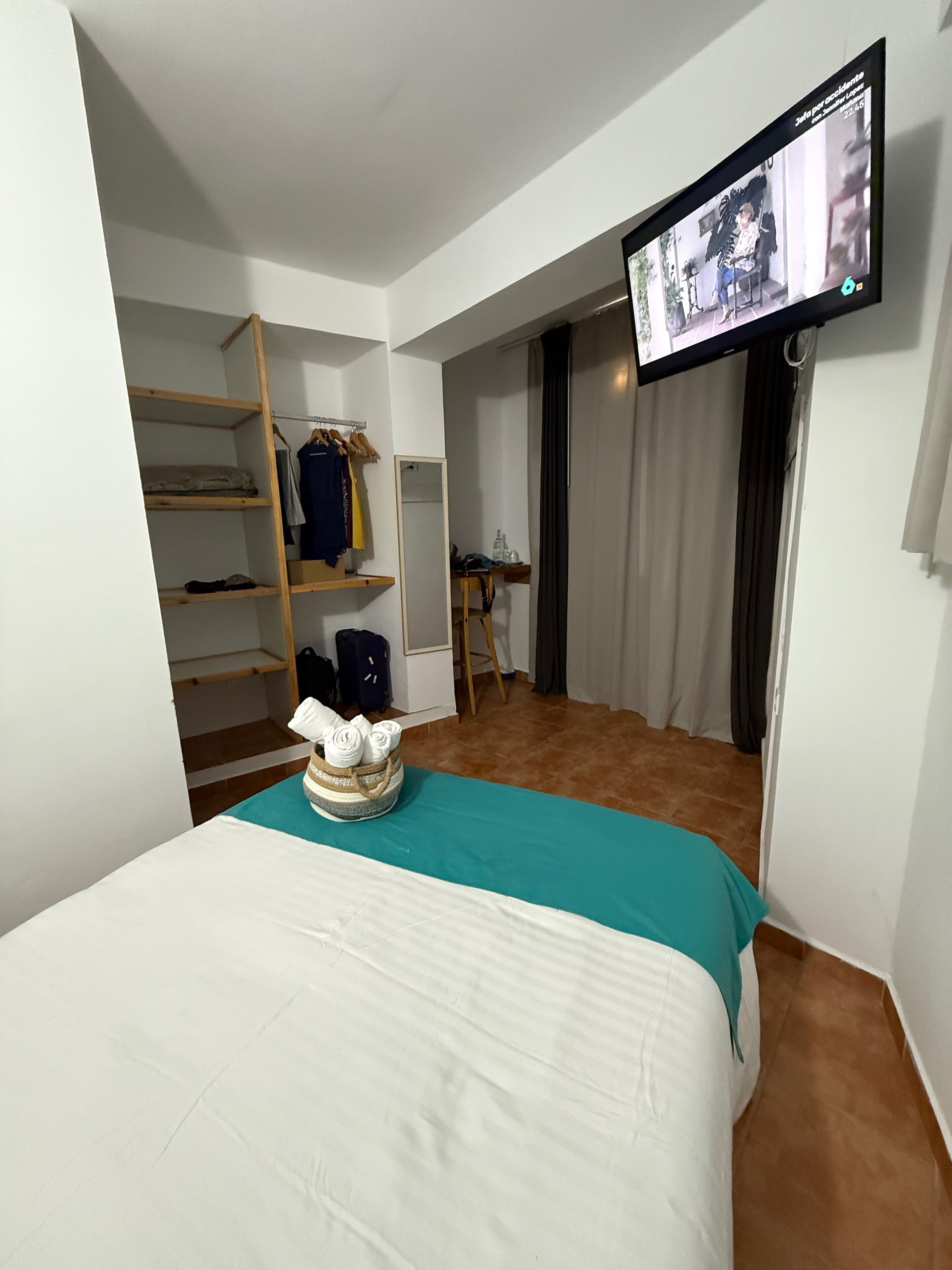 A hotel room with a bed, a television mounted on the wall, a wardrobe, and a small table with a chair.