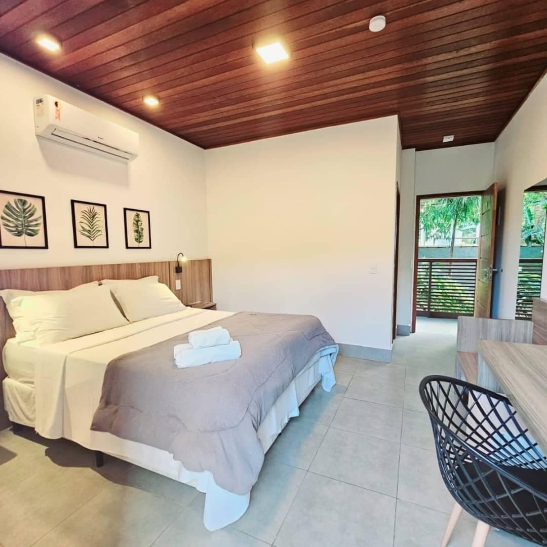 Een moderne slaapkamer met een houten plafond, een bed met een grijs-witte dekbedovertrek, een zwarte stoel, en een raam met uitzicht op groen.