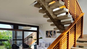 Una escalera moderna con barandas de madera y escalones de concreto, que conduce a una sala de estar con sofá, sillones y vistas al exterior.