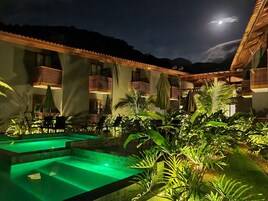 A nighttime view of a hotel with a pool area, outdoor seating, and a mountain backdrop.