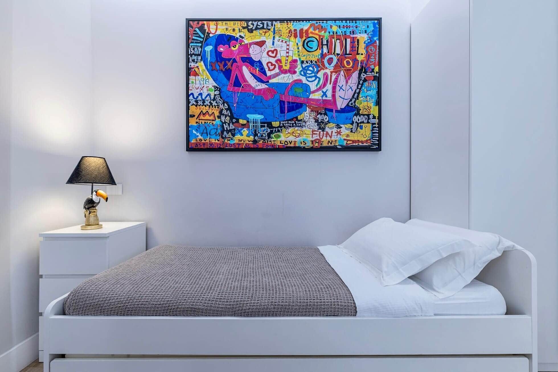 A bedroom with a white bed, a gray textured blanket, a white nightstand with a lamp, and a colorful abstract painting on the wall.