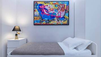A bedroom with a white bed, a gray textured blanket, a white nightstand with a lamp, and a colorful abstract painting on the wall.