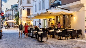 Een terras met tafels en stoelen, een geplaveid straatje en gebouwen met balkons.
