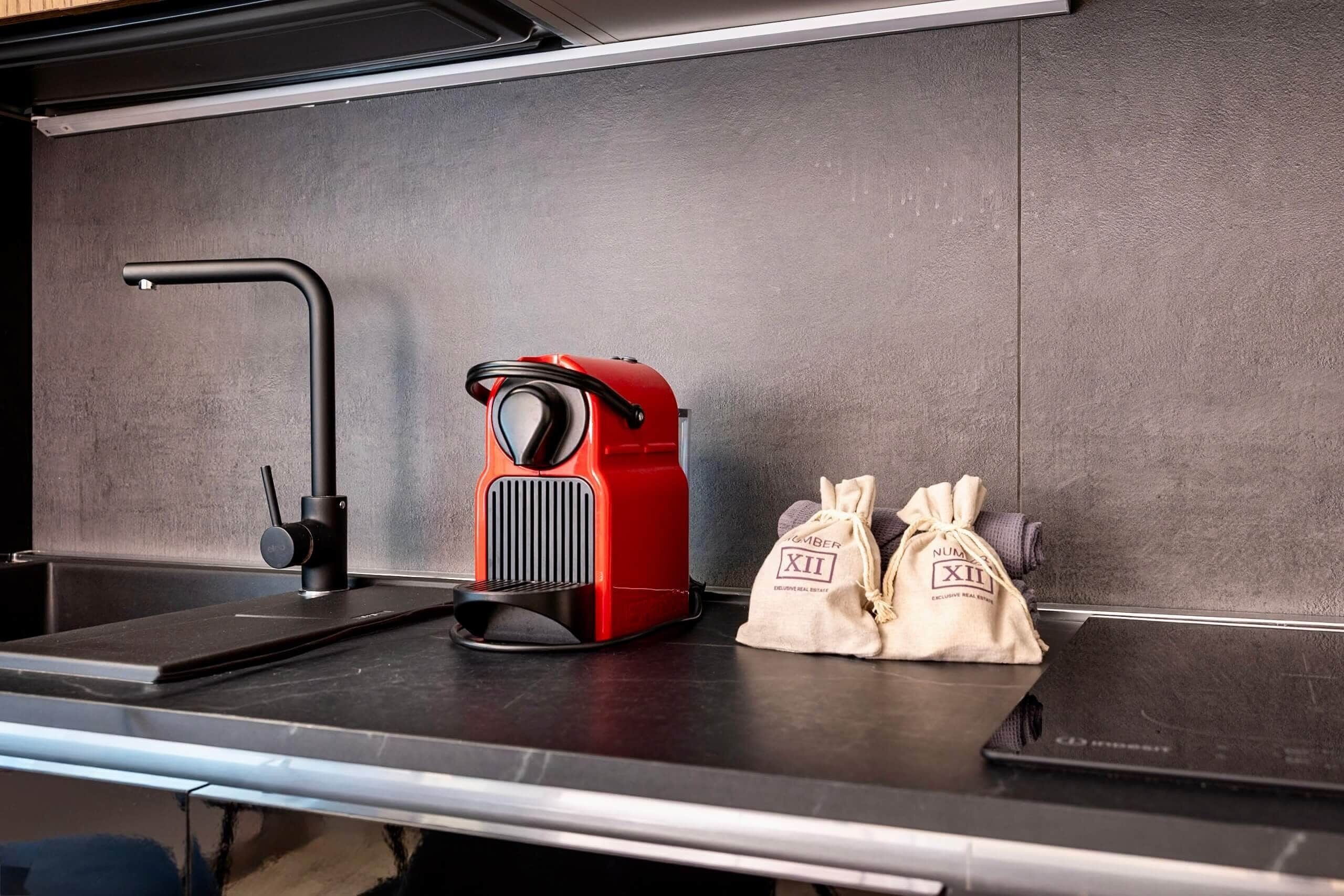 Un comptoir de cuisine avec un robinet moderne, une machine à café rouge et deux petits sacs étiquetés « NUMBERS XII ».