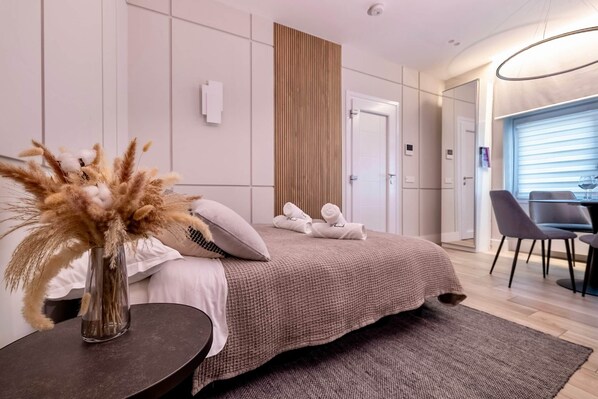 A modern hotel room with a large bed, a bedside table with a vase of dried flowers, a wooden panel wall, and a view of the dining area.