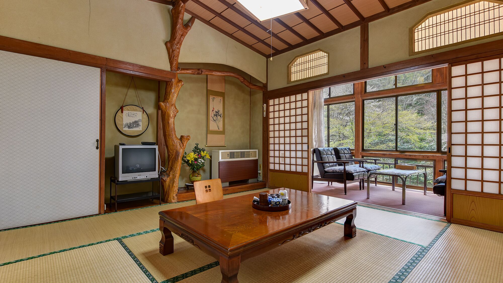 Egy hagyományos japán szoba, tatami szőnyeggel, alacsony asztallal, kanapéval és tolóajtókkal, melyekből zöld tájra nyílik kilátás.